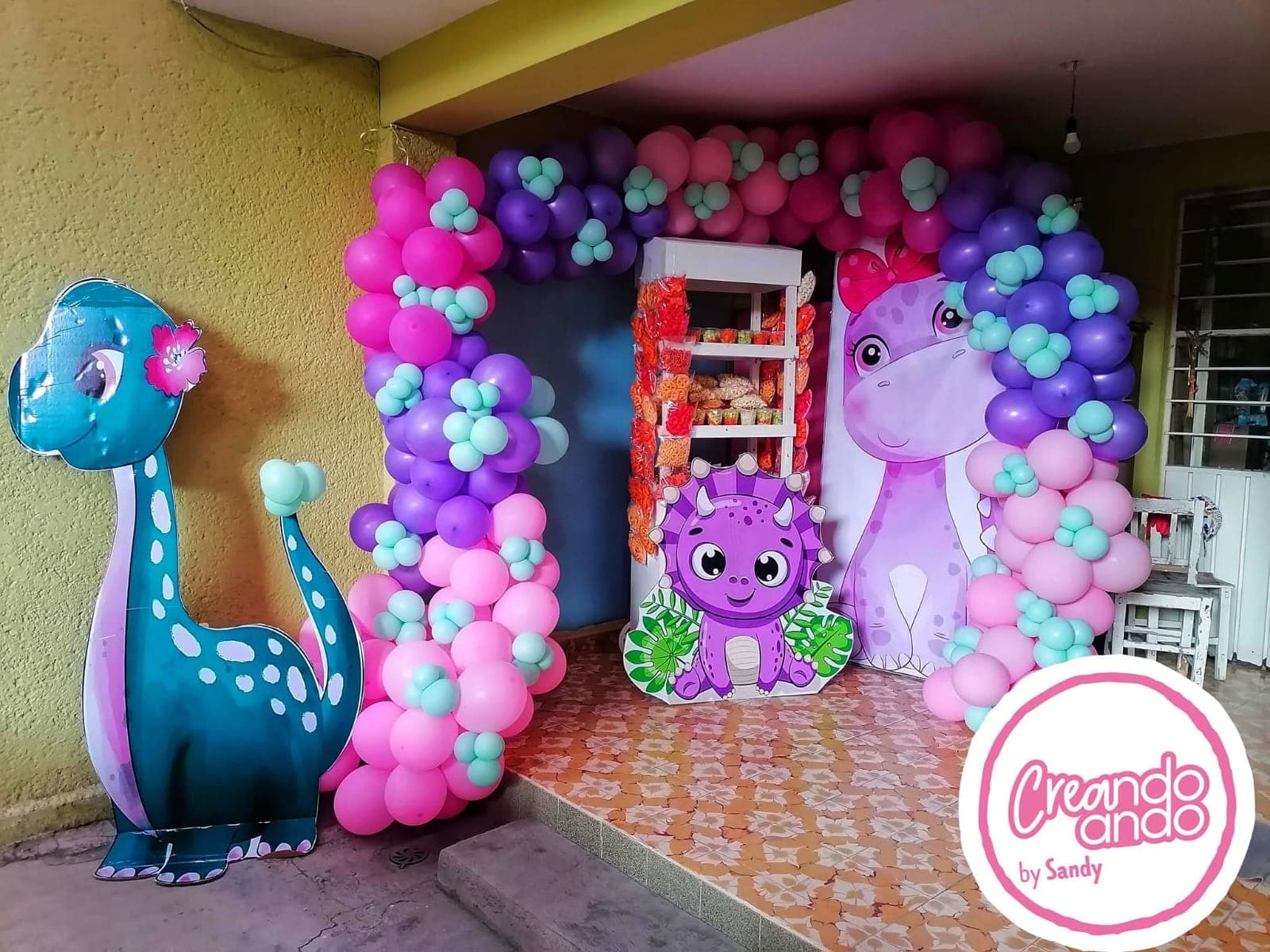 Estante blanco con bolsitas de botana dentro de un arco de globos rosa, morado y menta, con figuras de dinosaurio